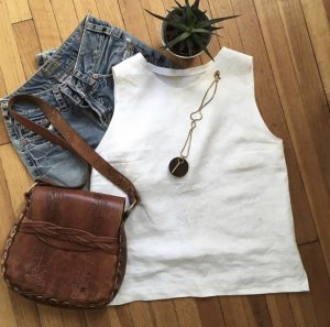 A flat-lay shot of a white, sleeveless shirt made by Heather Case and some thrifted clothing items.