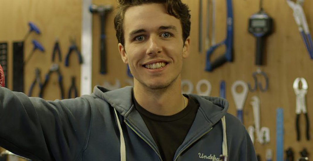 A man in a grey hoodie standing in front of a wall of tools.