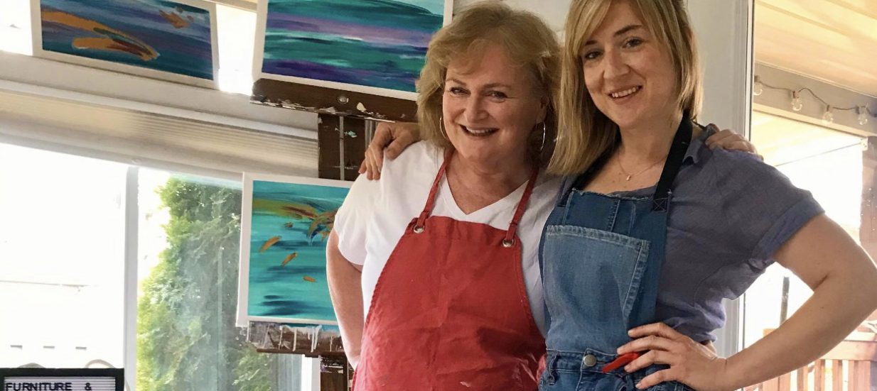 Two woman standing with their arms around each other, standing in front of landscape paintings.