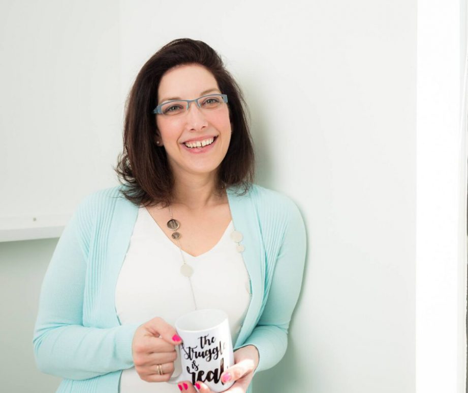 A person smiling and standing in front of a white wall.