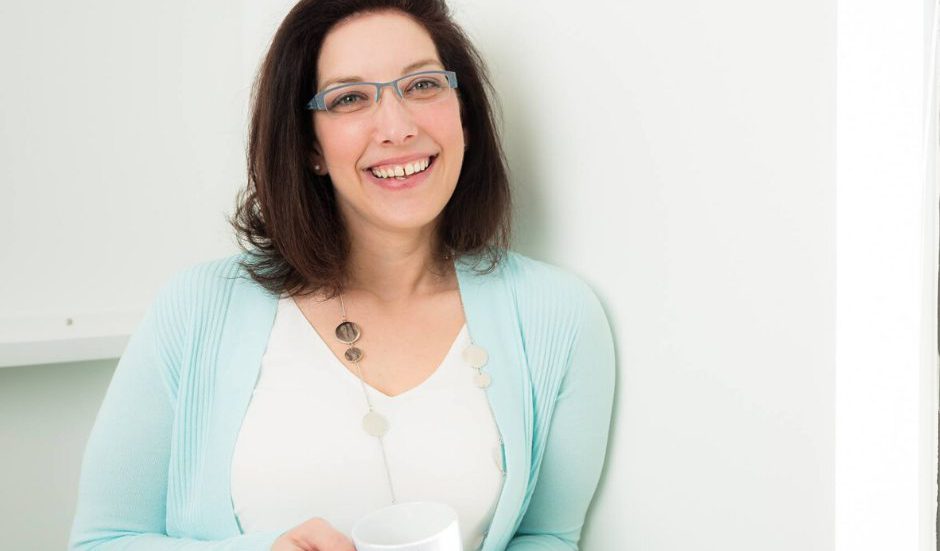 A person smiling and standing in front of a white wall.