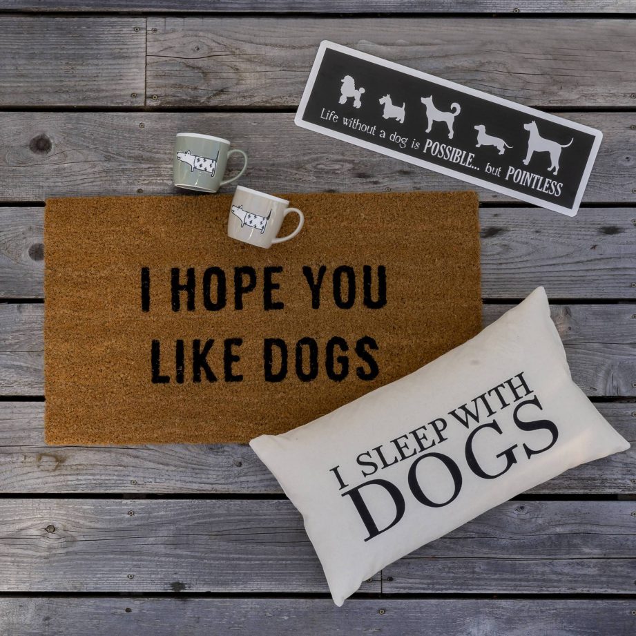 A door mat that says 'I hope you like dogs', a pillow that says 'I sleep with dogs' and two mugs on a wood background.