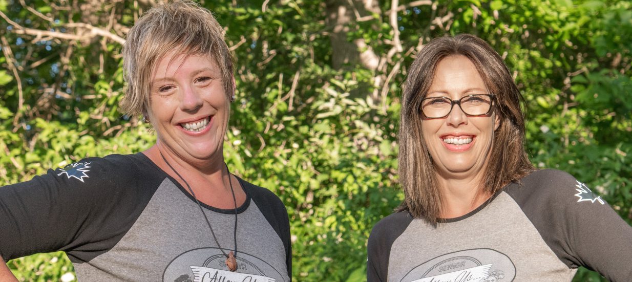 Two people standing side by side, in front of trees, smiling.