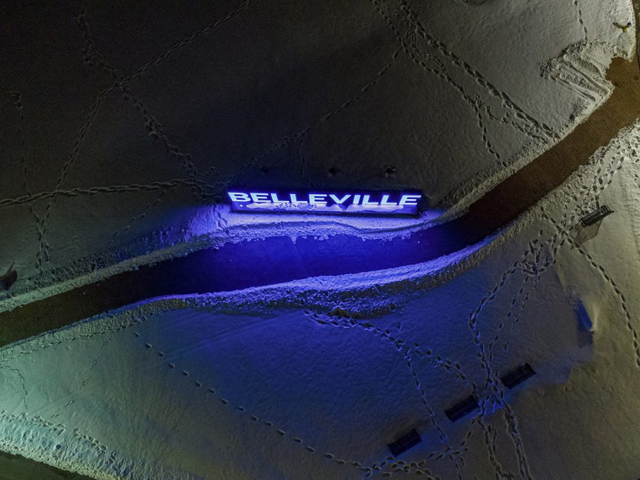 Aerial shot of snowy ground with footprint and a glowing 'Belleville' sign.