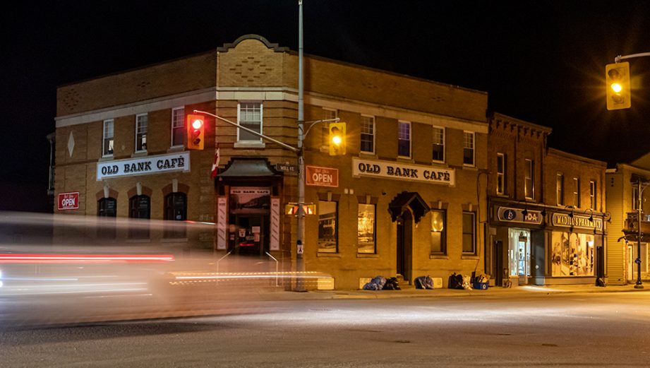 A street intersection at night with blurred cars. A building with a sign: Old Bank Cafe.