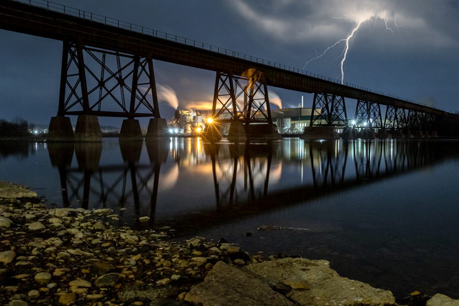 A lightning bolt at night behind a tall bridge crossing a river.