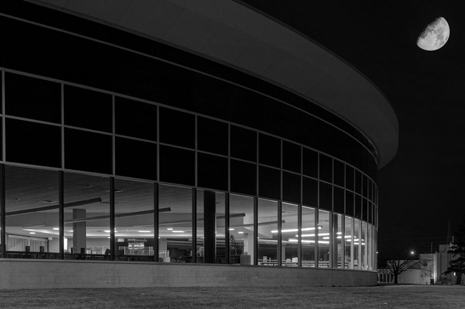 A black and white photo of a curved building with many windows, and the moon high in the sky.