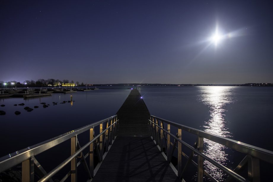 A boardwalk along the water at night with a bright moon in the sky.