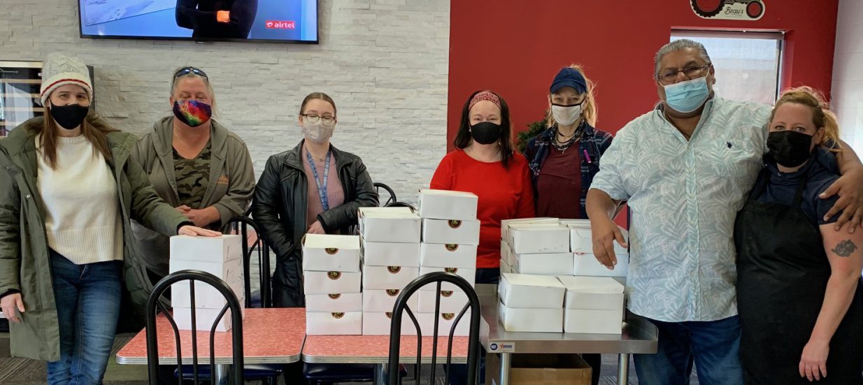 Seven masked people standing behind a table piled with takeout boxes.