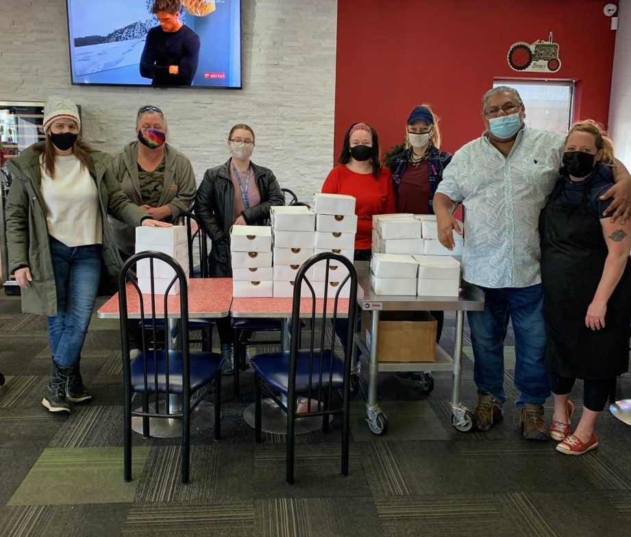 Seven masked people standing behind a table piled with takeout boxes.