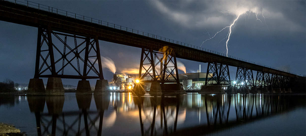 A lightning bolt at night behind a tall bridge crossing a river.