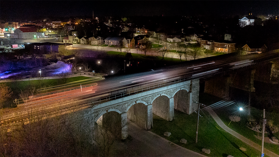 Aerial view of a stone bridge with arches at night, with a starry sky in the background and city lights.