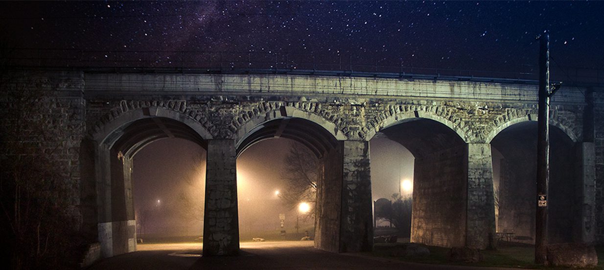 A stone bridge with arches at night, with a starry sky in the background.