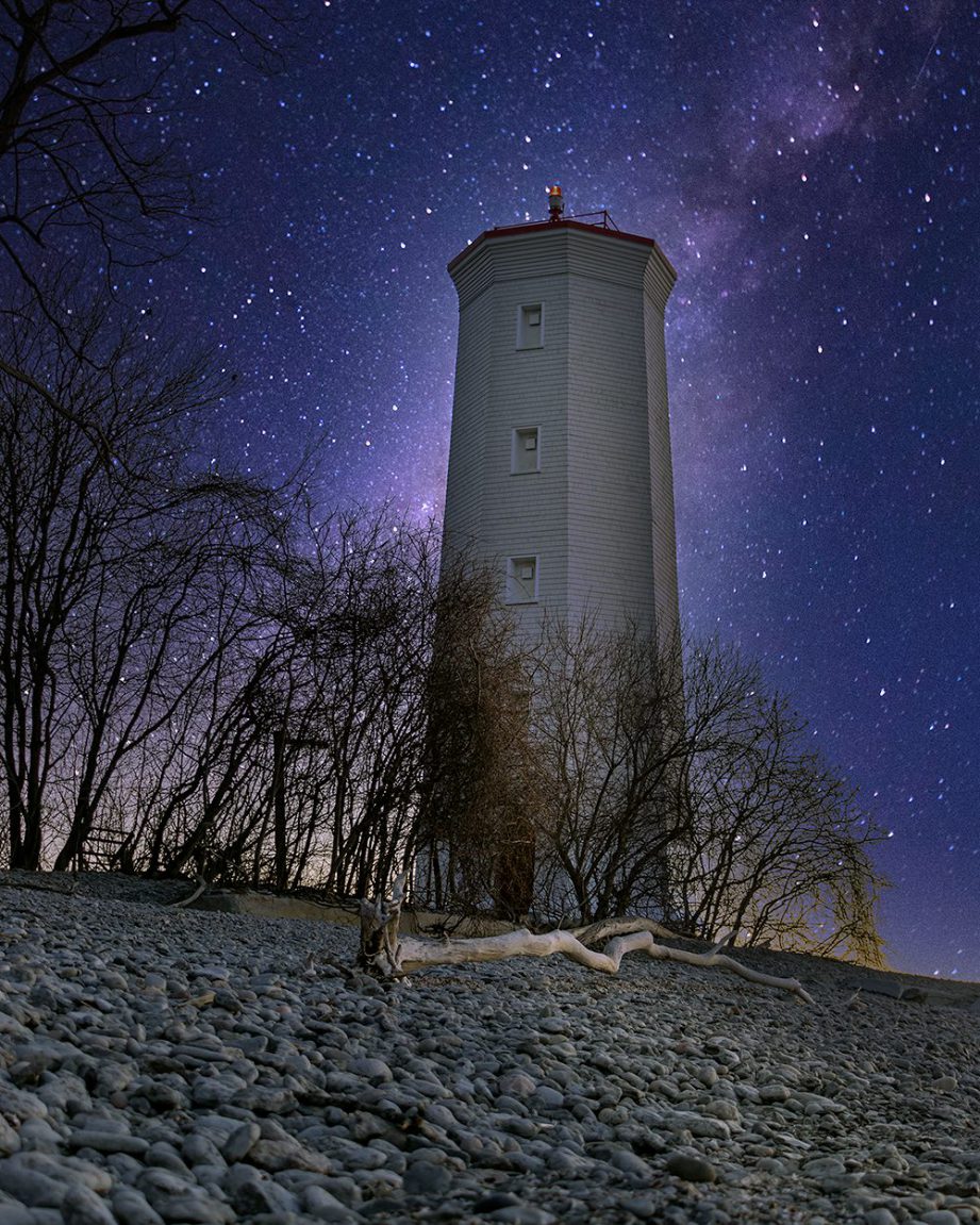 A lighthouse at night, along a stony beach with a starry sky in the background.