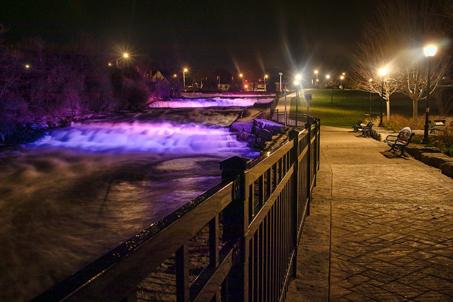 A long exposure of a river flowing, glowing with lights from the city, and a paved path alongside it.