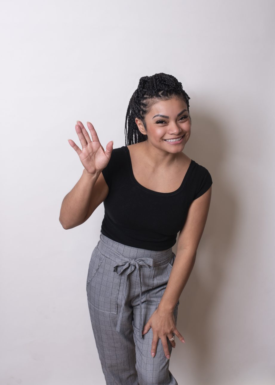 A person waving and smiling at the camera, standing in front of a white wall.