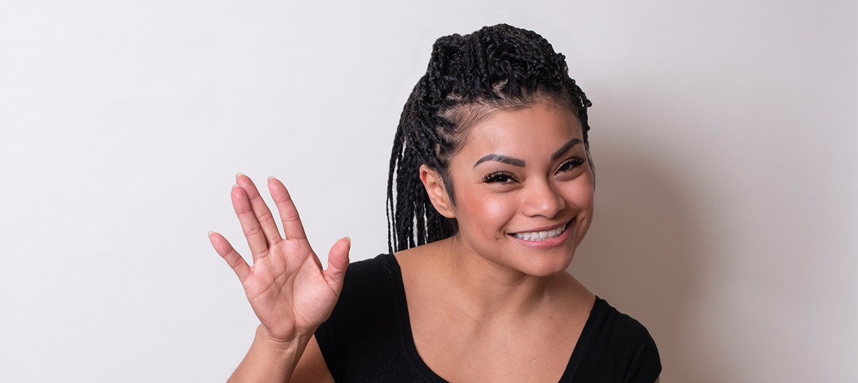 A person waving and smiling at the camera, standing in front of a white wall.