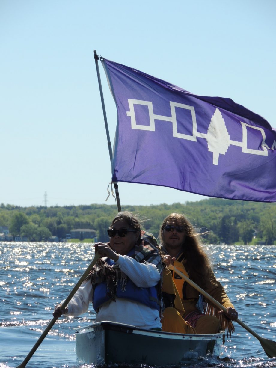 Two people in a canoe paddling in the water, with the purple Haudenosaunee flag.