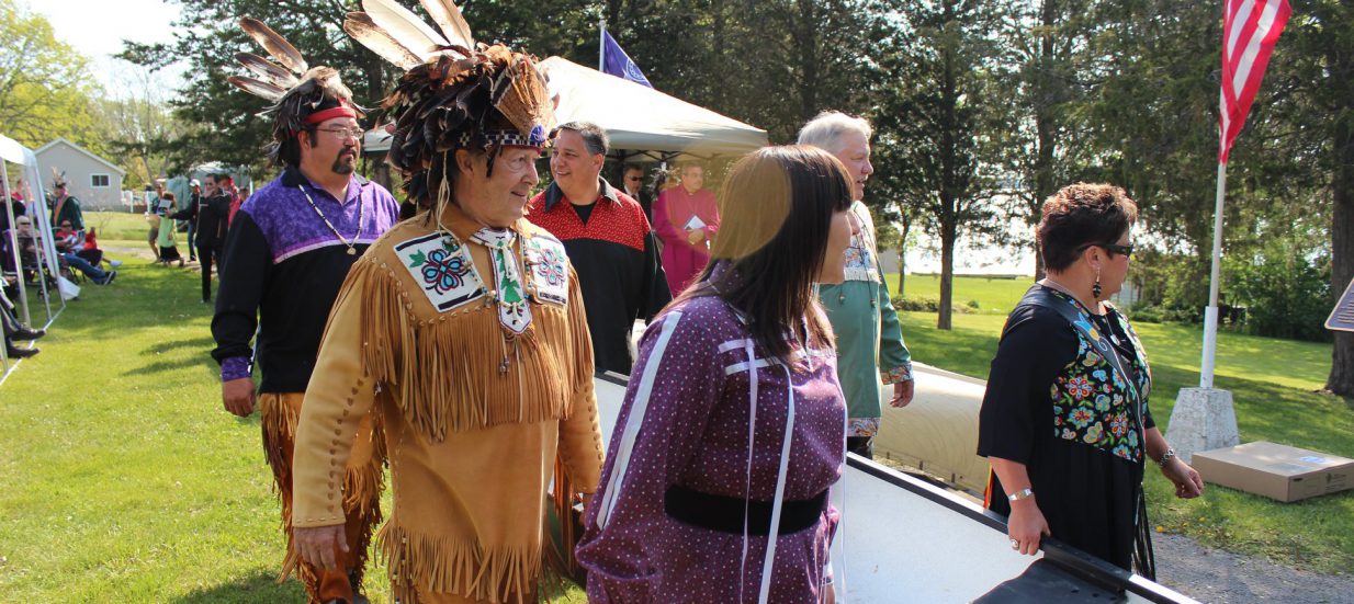 a group of Mohawk people dressed in regalia holding a canoe and walking on grass towards the water for the Mohawk Landing celebration.