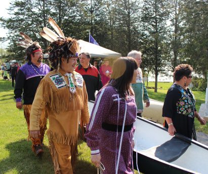 a group of Mohawk people dressed in regalia holding a canoe and walking on grass towards the water for the Mohawk Landing celebration.