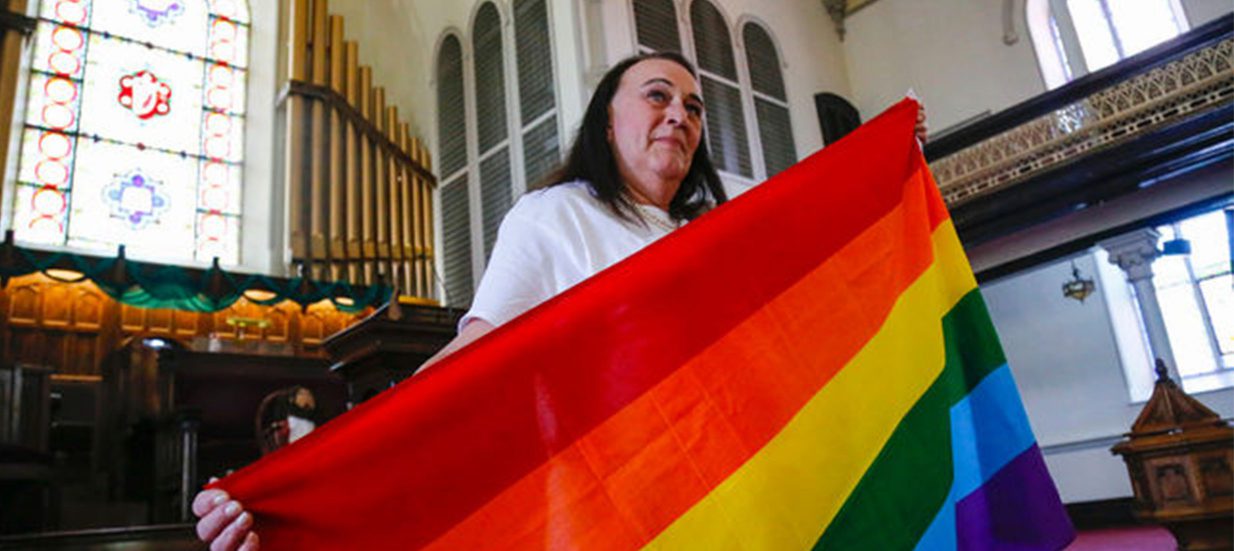 A person wearing a white shirt holding a rainbow flag in a church.