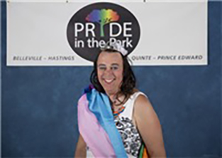 A person wearing a crown and sash, standing in front of a white sign with text: PRIDE in the Park.