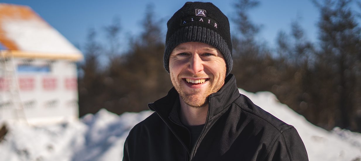 A person wearing a toque and winter jacket smiling, with snow and a partially constructed house in the background.