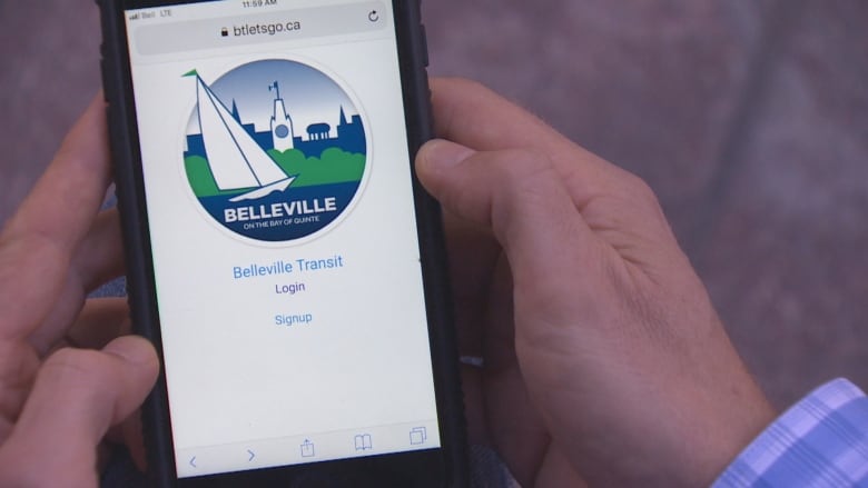 Two hands holding a phone. On the screen there is a white background, logo for the City of Belleville and text below: Belleville Transit, log in or sign up.