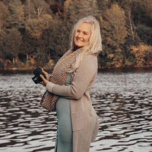 A person holding a camera standing by water with a forest in the background.