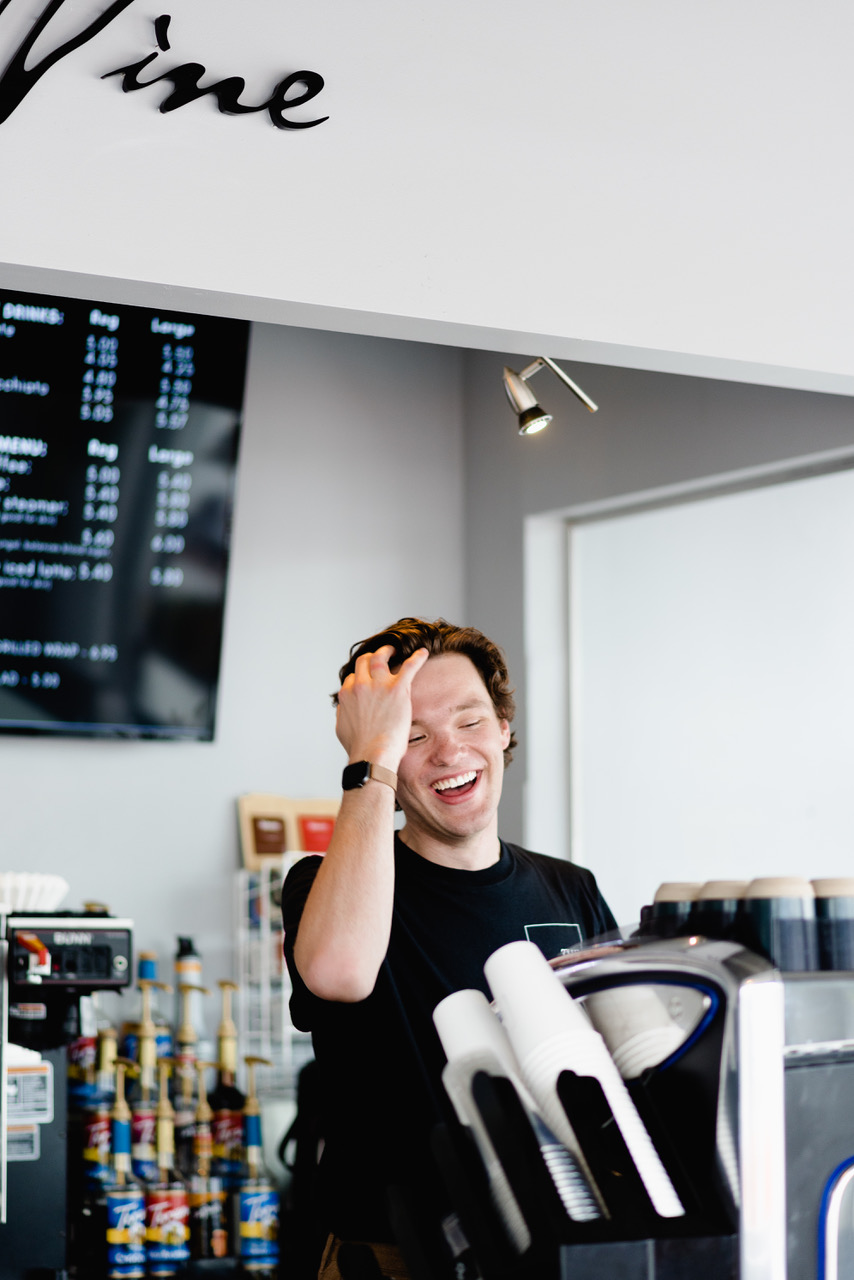 A person standing behind the counter at a cafe, laughing and running a hand through his hair.