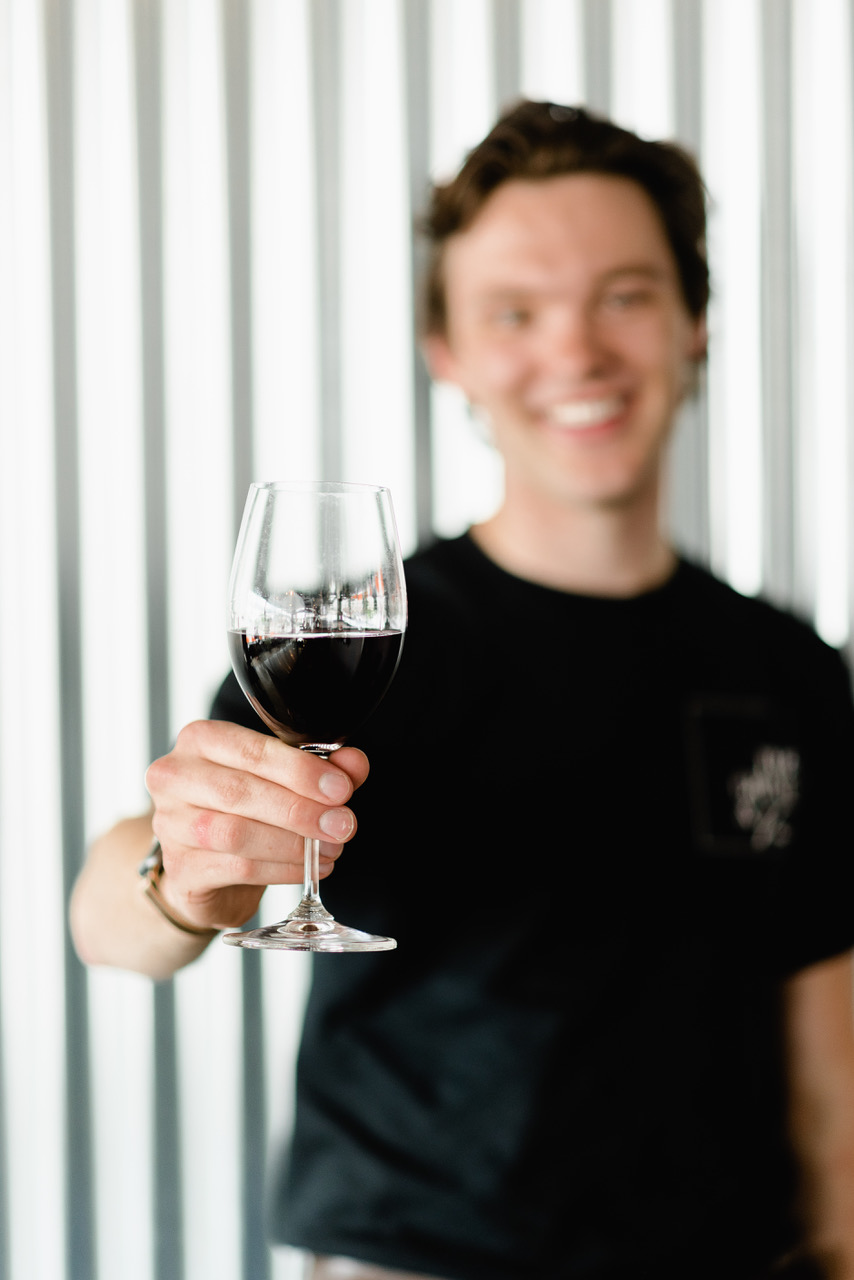 A blurred photo of a person wearing a black shirt, holding a wine glass which is in focus.