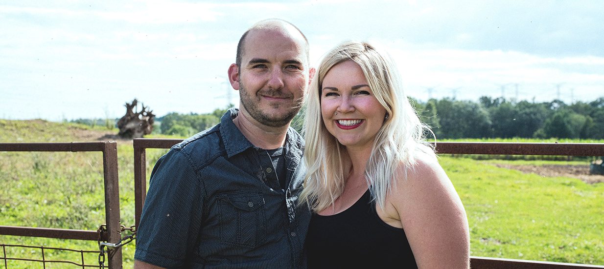 Brad and Rachel Gibson, farmers and owners of Opoma Farms in Quinte West, standing side by side in front of a field.