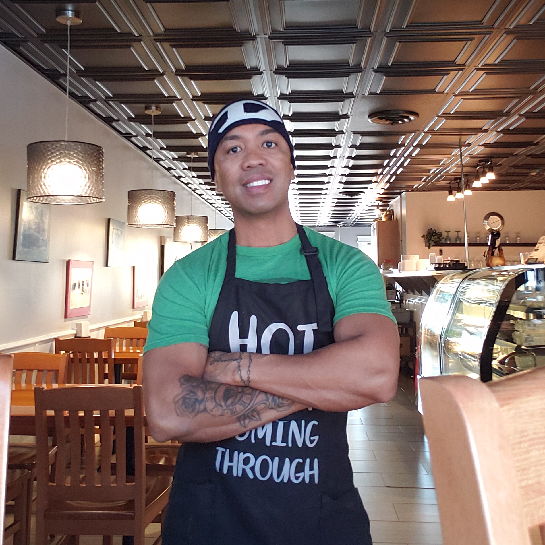 A person wearing a green shirt and black apron standing in a cafe.