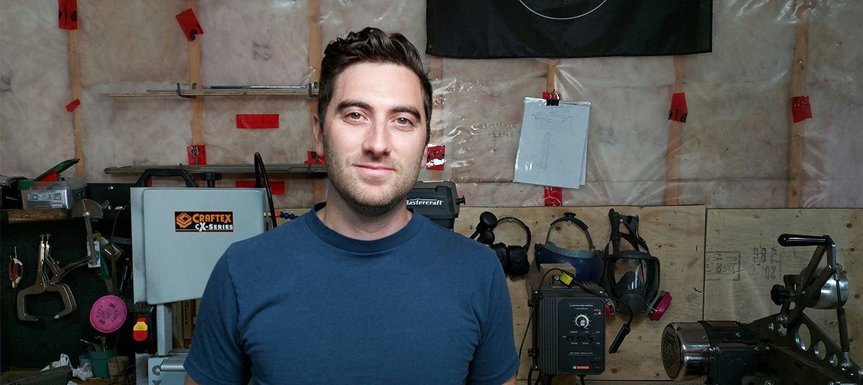 A person wearing a blue shirt standing in a garage with tools behind him.