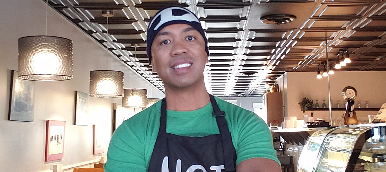 A person wearing a green shirt and black apron standing in a cafe.