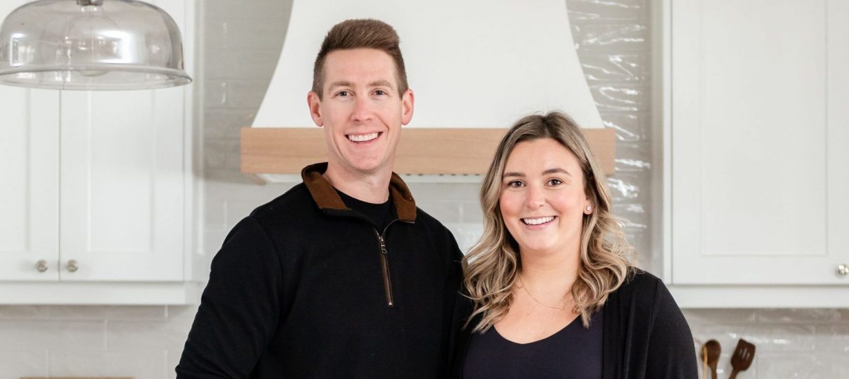 Andrew and Monique Guthrie of Park Road Design & Build posed together in a newly-built kitchen with white cabinets, an island and marble countertops.