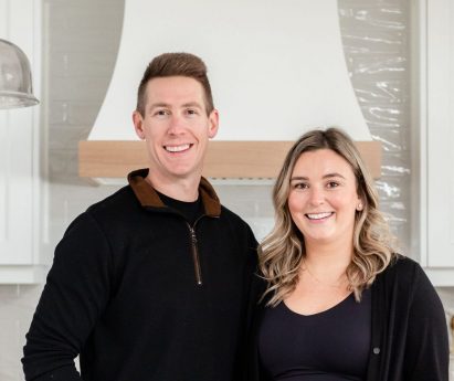 Andrew and Monique Guthrie of Park Road Design & Build posed together in a newly-built kitchen with white cabinets, an island and marble countertops.