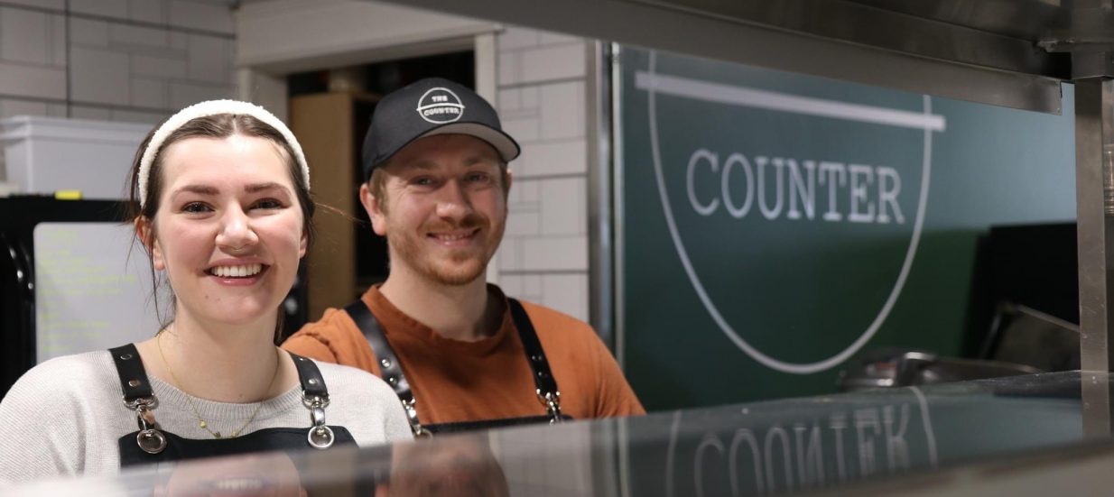 Kylie and Braden Lawther, owners of The Counter in downtown trenton, pose behind the counter to their kitchen in front of their sign.