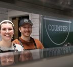 Kylie and Braden Lawther, owners of The Counter in downtown trenton, pose behind the counter to their kitchen in front of their sign.