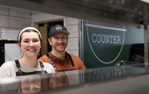 Kylie and Braden Lawther, owners of The Counter in downtown trenton, pose behind the counter to their kitchen in front of their sign.