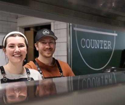 Kylie and Braden Lawther, owners of The Counter in downtown trenton, pose behind the counter to their kitchen in front of their sign.