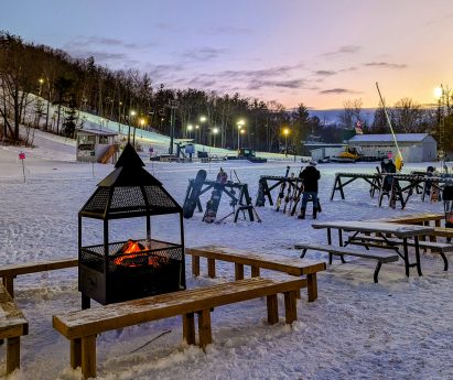A firepit with benches in front of the chalet at the bottom of the slopes at Batawa Ski Hill. There are racks of skis and snowboards and the slopes are in the background.