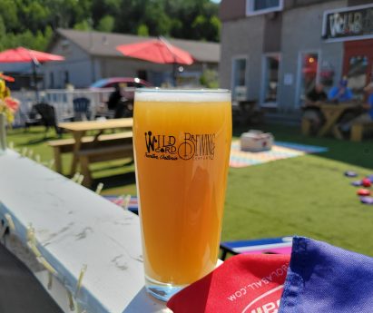 a pint of a hazy IPA beer in a glass with the Wild Card Brewing Co logo sits on the railing of the Wild Card Brewing Company patio on a sunny day. Lawn games and picnic tables in the background